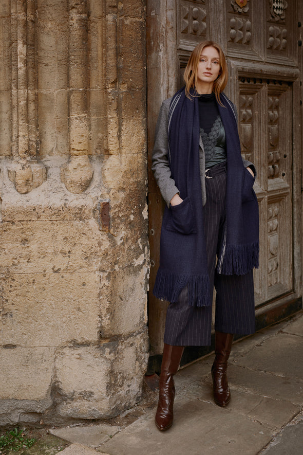 Model wearing large blanket wrap in indigo Merino wool and cream edge detail hands in the patch pockets