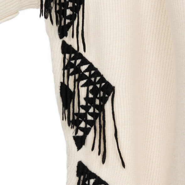 Close up of embroidered edge of one size poncho loose-fitting wrap with sleeves knitted in cream 50% cashmere/wool mix edged with a hand-embroidered geometric design in charcoal grey black with trailing threads along edge of body and sleeves from Thread Tales company